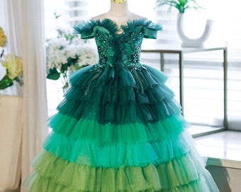 May include: A formal gown in shades of green, displayed on a mannequin. The off-the-shoulder bodice is embellished with intricate beadwork. The skirt features layers of ruffled fabric, transitioning from dark to light green.