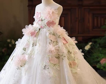 May include: A floor-length white dress featuring a sleeveless bodice and a full, voluminous skirt. The garment is adorned with layered, three-dimensional fabric flowers in shades of soft pink and white, accented with green leaf embroidery.