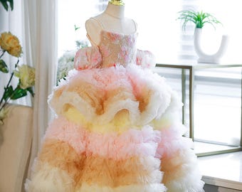 May include: A multi-layered, pastel-colored dress on a mannequin. The bodice is embellished with floral details and the skirt features layers of pink, yellow, and white ruffles. The dress has a square neckline and off-the-shoulder sleeves.