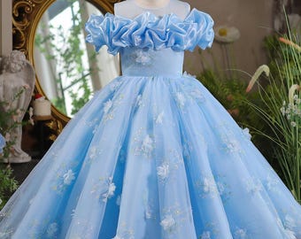 May include: A light blue formal dress with a tulle skirt and floral embellishments. The bodice features ruffled sleeves and a sheer neckline. The dress is displayed on a mannequin, showcasing its elegant design and details.