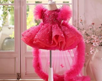 May include: A bright pink dress with a layered skirt and feathered details on the shoulders and train. The dress is displayed on a white mannequin stand, highlighting its intricate design and texture. Ideal for a special event.