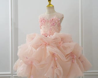 May include: A light pink, layered tulle dress with floral embellishments. The bodice features delicate pink flowers and the skirt has multiple layers of soft tulle. The dress is displayed on a mannequin.