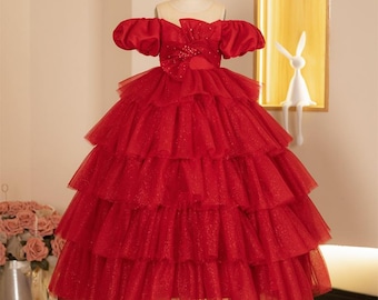 May include: A red formal dress with a layered skirt and off-the-shoulder sleeves. The bodice features a bow detail and the dress is displayed on a mannequin. The dress is made of a shimmering fabric.