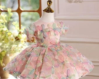 May include: A light pink dress with floral embellishments. The dress features a round neckline and short sleeves, adorned with pastel-colored flowers. The skirt is full and layered, creating a voluminous silhouette. The dress is displayed on a mannequin.