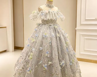 May include: A floor-length gown in a light gray hue featuring an off-the-shoulder neckline with ruffled sleeves. The bodice and skirt are adorned with silver sequins and three-dimensional floral appliqués in soft shades of blue and pale green.