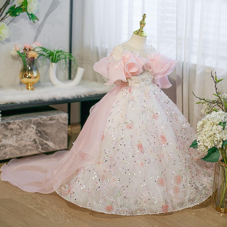 May include: A white and pink formal dress with a long train. The bodice has pink ruffled sleeves and a floral design. The skirt is decorated with sequins and pink floral accents. The dress is displayed on a mannequin.