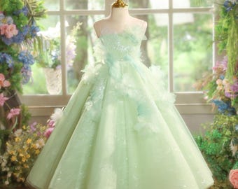 May include: A pale green formal dress on a mannequin. The dress has a tulle skirt and bodice with floral accents and sequins. The dress is displayed in front of a window with flowers.