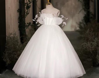 May include: A white, strapless dress with a full tulle skirt. The bodice is adorned with white feathers and floral accents. The dress is displayed on a mannequin with a gold-colored headpiece, showcasing its elegant design.