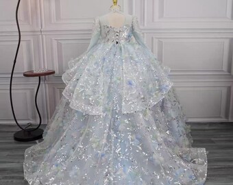 May include: A floor-length light blue ball gown featuring long sleeves, a high neckline, and a corset-style lace-up back. The dress is adorned with silver sequins and scattered blue and white floral appliques, creating a voluminous skirt.