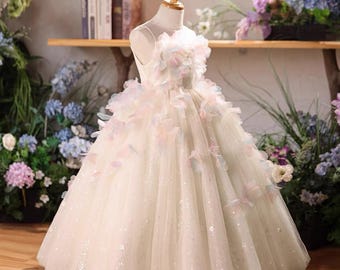 May include: A floor-length sleeveless dress in a cream hue, featuring a bodice and skirt adorned with layered, multi-colored pastel fabric petals. The skirt is crafted from layers of tulle with a subtle shimmer, creating a voluminous silhouette.