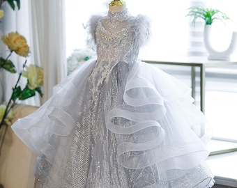 May include: A silver and white formal dress with a high neckline and layered skirt. The bodice features intricate beading and lace details, with feather accents at the shoulders. The dress is displayed on a mannequin.