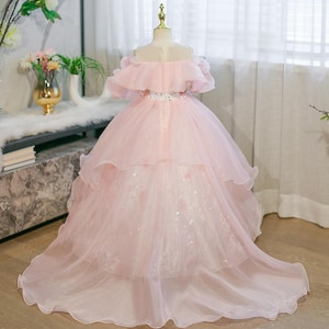 May include: A pale pink off-the-shoulder dress with a layered skirt and a long train. The dress has a ruffled neckline and a beaded belt. The skirt features multiple layers of tulle and a flowing train, creating a formal appearance.