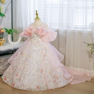 May include: A formal, floor-length dress in white and pink, designed for a young girl. The dress features a bodice with sparkling embellishments and off-the-shoulder, ruffled sleeves. The skirt is layered with a flowing train, adorned with floral accents and sequins.