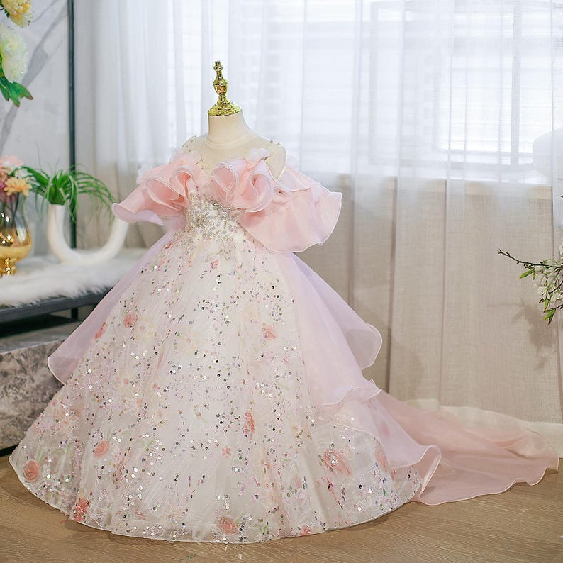 May include: A formal, floor-length dress in white and pink, designed for a young girl. The dress features a bodice with sparkling embellishments and off-the-shoulder, ruffled sleeves. The skirt is layered with a flowing train, adorned with floral accents and sequins.