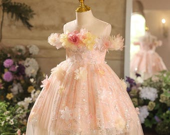 May include: A light pink, off-the-shoulder dress on a mannequin. The dress is adorned with floral embellishments in shades of pink, yellow, and white. The skirt has a layered design with a shimmering, sequined bodice. The dress is designed for a special occasion.