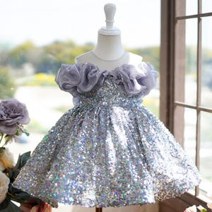 May include: A silver sequined dress with a lilac ruffled neckline and bodice, displayed on a mannequin. The dress has a flared skirt and is designed for a child. The background includes a window and floral arrangements.