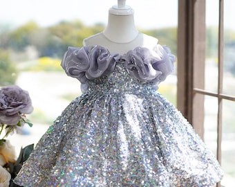 May include: A silver sequined dress with a lilac ruffled neckline and bodice, displayed on a mannequin. The dress has a flared skirt and is designed for a child. The background includes a window and floral arrangements.
