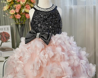May include: A formal dress with a black sequined bodice and a large black bow. The skirt is a light pink with layers of petal-like fabric. The neckline is embellished with sparkling stones. The dress is displayed on a mannequin.