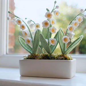 以下が含まれることがあります： 黄色い中心部を持つ白い鐘形の複数の花と緑の葉が特徴の、かぎ針編みのスズランの植物。茎の根元に緑色の苔が敷き詰められた、長方形の白い陶器製のプランターに植えられています。