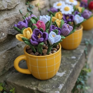May include: A yellow ceramic mug with a grid pattern, filled with small brown stones and a bouquet of crocheted flowers. The flowers are in shades of purple, white, yellow, and pink, with green leaves. The mug is sitting on a stone surface.