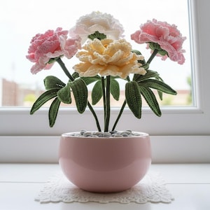 May include: A handmade crochet flower arrangement featuring pink, white, and yellow peonies. The flowers are in a pink ceramic pot filled with small gray stones. Green leaves and stems support the flowers, creating a decorative floral display.