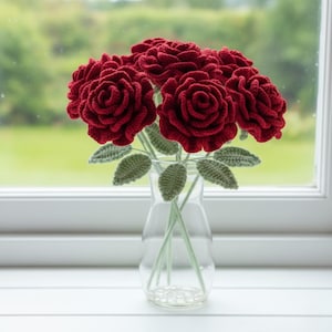 Puede incluir: Un jarrón de vidrio transparente contiene un ramo de rosas rojas hechas a mano y tejidas a ganchillo. Las rosas tienen pétalos detallados y se combinan con hojas y tallos verdes tejidos a ganchillo. El jarrón está sobre una superficie blanca, con un fondo verde borroso.