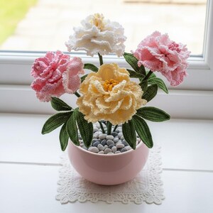 May include: A handcrafted crochet flower arrangement in a pink ceramic pot. The arrangement features five flowers in shades of pink, yellow, and white, with green leaves. The pot is filled with small gray stones and sits on a white lace doily.