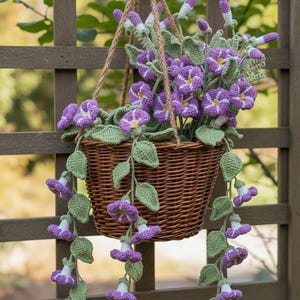 May include: A brown woven wicker hanging basket filled with crocheted purple morning glory flowers and green leaves. The flowers feature white centers and yellow stamens, with long trailing vines cascading down from the basket, suspended by jute rope.