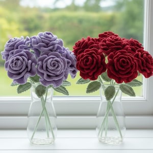 May include: Two vases of crochet roses sit on a windowsill. One vase holds purple roses, the other holds red roses. The roses have green stems and leaves. The vases are clear glass.