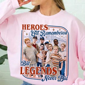 May include: Pink sweatshirt featuring a graphic of a group of boys and the text "HEROES Get Remembered But LEGENDS Never Die" in red and blue. The sweatshirt has a crew neck and long sleeves.