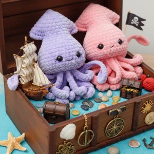 Peut inclure: Un coffre au trésor en bois rempli d'objets sur le thème nautique, dont deux calmars crochetés en lavande et rose. Un voilier miniature, des pièces de monnaie et un drapeau pirate complètent la scène. Le coffre est décoré de coquillages et d'une boussole.