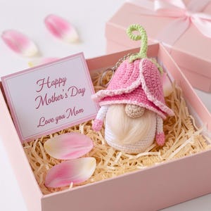 Puede incluir: Un llavero de gnomo de ganchillo hecho a mano con un sombrero de flor rosa, dentro de una caja de regalo rosa con una tarjeta que dice "Happy Mother's Day Love you Mom". La caja está llena de paja y decorada con pétalos de flores rosas.