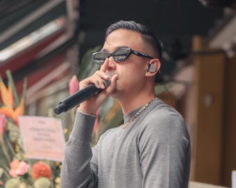 May include: A person wearing sunglasses and a gray long-sleeved shirt holds a microphone. They are wearing a gold chain necklace and in-ear monitors. The background includes flowers and a sign.