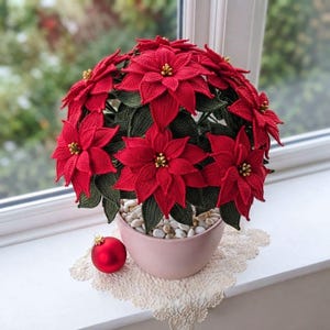 Op de afbeelding: Een gehaakte kerststerplant met levendige rode bloemblaadjes en donkergroene bladeren, voorzien van kleine gele hartjes. De plant staat in een gladde, lichtroze keramische pot gevuld met witte steentjes, vergezeld door een rode kerstbal.