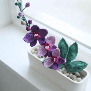 May include: A crocheted orchid plant featuring deep purple and light lavender petals with dark magenta centers. The plant has dark green leaves and a long stem with small buds, all potted in a rectangular white container filled with smooth white stones.