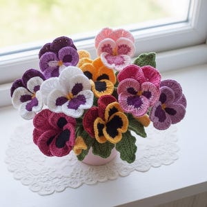 May include: A crochet arrangement of pansies in a small pink pot. The flowers are in shades of purple, white, orange, red, and pink, with green leaves. The bouquet rests on a white surface with a lace doily.