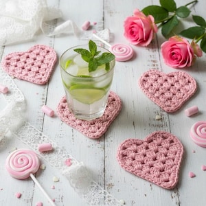 Puede incluir: Una bebida refrescante con hielo y lima descansa sobre un posavasos rosa en forma de corazón tejido. La imagen también presenta otros tres posavasos de corazón rosa, rosas rosas, una piruleta y encaje decorativo sobre una superficie de madera blanca.