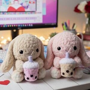 Puede incluir: Dos conejos de peluche, uno beige y otro rosa, cada uno sosteniendo un té boba de ganchillo. Las tazas de té boba son rosas y beige con bobas negras y una pajita morada. Los conejos tienen grandes ojos negros y largas orejas caídas.