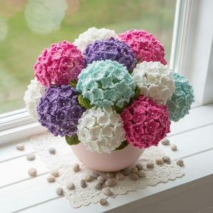 May include: A bouquet of crocheted hydrangea flowers in shades of pink, purple, white, and light blue, each featuring small white beads at the center of the petals. The flowers are arranged in a rounded pink bowl with green crocheted leaves.