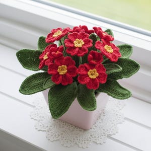 May include: A handmade crochet flower arrangement featuring vibrant red flowers with yellow centers and green leaves. The flowers are in a light pink square pot, sitting on a white lace doily. The arrangement is placed on a white windowsill.