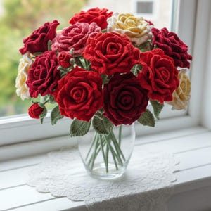 Op de afbeelding: Een boeket gehaakte rozen in een heldere glazen vaas. De rozen zijn in verschillende tinten rood en geel, met groene bladeren en stelen. De vaas staat op een witte vensterbank met een kanten kleedje.