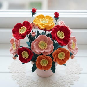 Puede incluir: Un arreglo floral hecho a mano con ganchillo en una maceta rosa. Las flores son en tonos de rojo, naranja, amarillo y rosa, con hojas verdes. Las flores tienen centros amarillos. El arreglo está sobre un tapete de encaje blanco.
