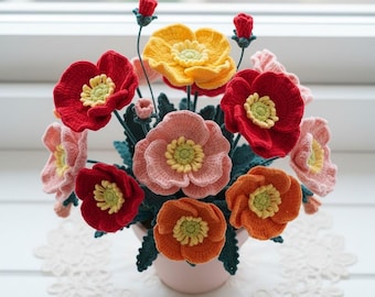 May include: A handmade crochet flower arrangement in a pink pot. The flowers are in shades of red, orange, yellow, and pink, with green leaves. The flowers have yellow centres. The arrangement is on a white lace doily.