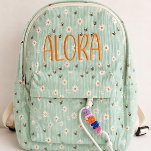 Puede incluir: Una mochila de pana verde claro con un patrón repetitivo de margaritas blancas y pequeñas abejas. El nombre ALORA está bordado en hilo naranja en la parte frontal. Un tirador de cremallera de cordón blanco muestra el nombre ALORA en cuentas.
