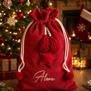 Könnte beinhalten: Ein roter Geschenksack aus Kord mit Kordelzug, verziert mit dem in Weiß gestickten Namen Alora, einem zentralen roten Stern-Applikation sowie zwei weißen Kordeln mit Holzperlen, roten Quasten und roten Bommeln an den Enden.