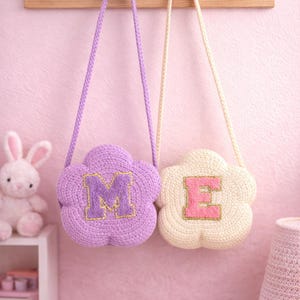 Puede incluir: Dos bolsos de ganchillo con forma de flor y correas largas cuelgan de un perchero de madera. Uno es lavanda con una "M" con ribete dorado, el otro es crema con una "E" con ribete rosa. Los bolsos están en una habitación rosa.