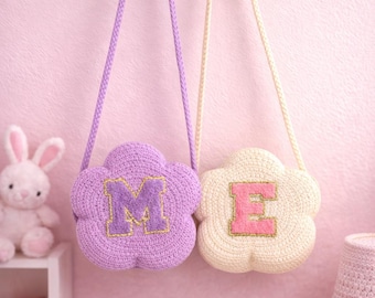 May include: Two flower-shaped crochet bags with long straps hanging from a wooden rack. The purple bag features a large letter M in a darker purple, and the cream-colored bag features a large letter E in pink. Both letters have gold stitching.