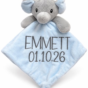 May include: A soft, light blue plush security blanket featuring a gray elephant head with light blue inner ears. The blanket is embroidered with the name EMMETT and the date 01.10.26 in dark gray thread, centered on the diamond-shaped fabric.