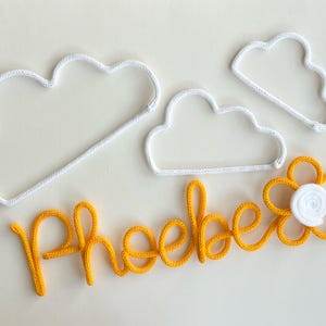 May include: Handmade nursery decoration. The image shows three white crochet cloud shapes, the name "Phoebe" in yellow yarn, and a yellow flower with a white centre. The items are arranged on a light background.