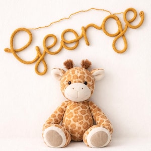 May include: A soft giraffe toy with a brown and tan spotted pattern, positioned beneath a mustard yellow yarn name, "GEORGE". The name is written in a cursive style, perfect for nursery decoration.